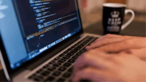 Close-up of hands typing on a laptop with code on the screen and a coffee cup beside it.