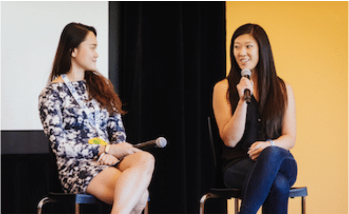 Two women sit on stage during a panel discussion, holding microphones, with an engaging backdrop in a professional setting.