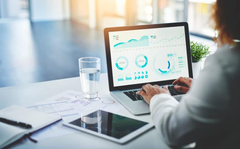 Person at a desk analyzing graphs on a laptop screen with documents and a tablet nearby.