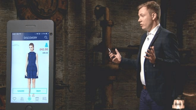 A presenter showcases an app featuring a blue dress priced at £42 on a smartphone in a business setting.