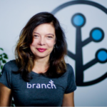 Branch Co-founder Mada Seghete smiling at camera in a Branch t-shirt in front of the Branch logo glyph