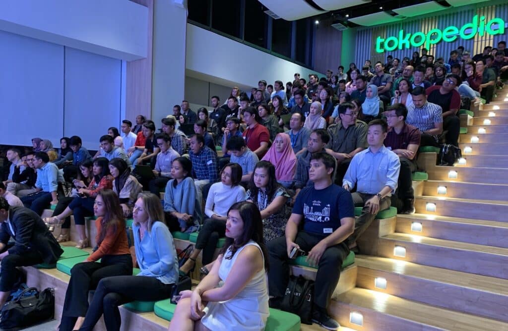 Audience seated on tiered steps in a well-lit venue with a large "tokopedia" sign in the background during an event.