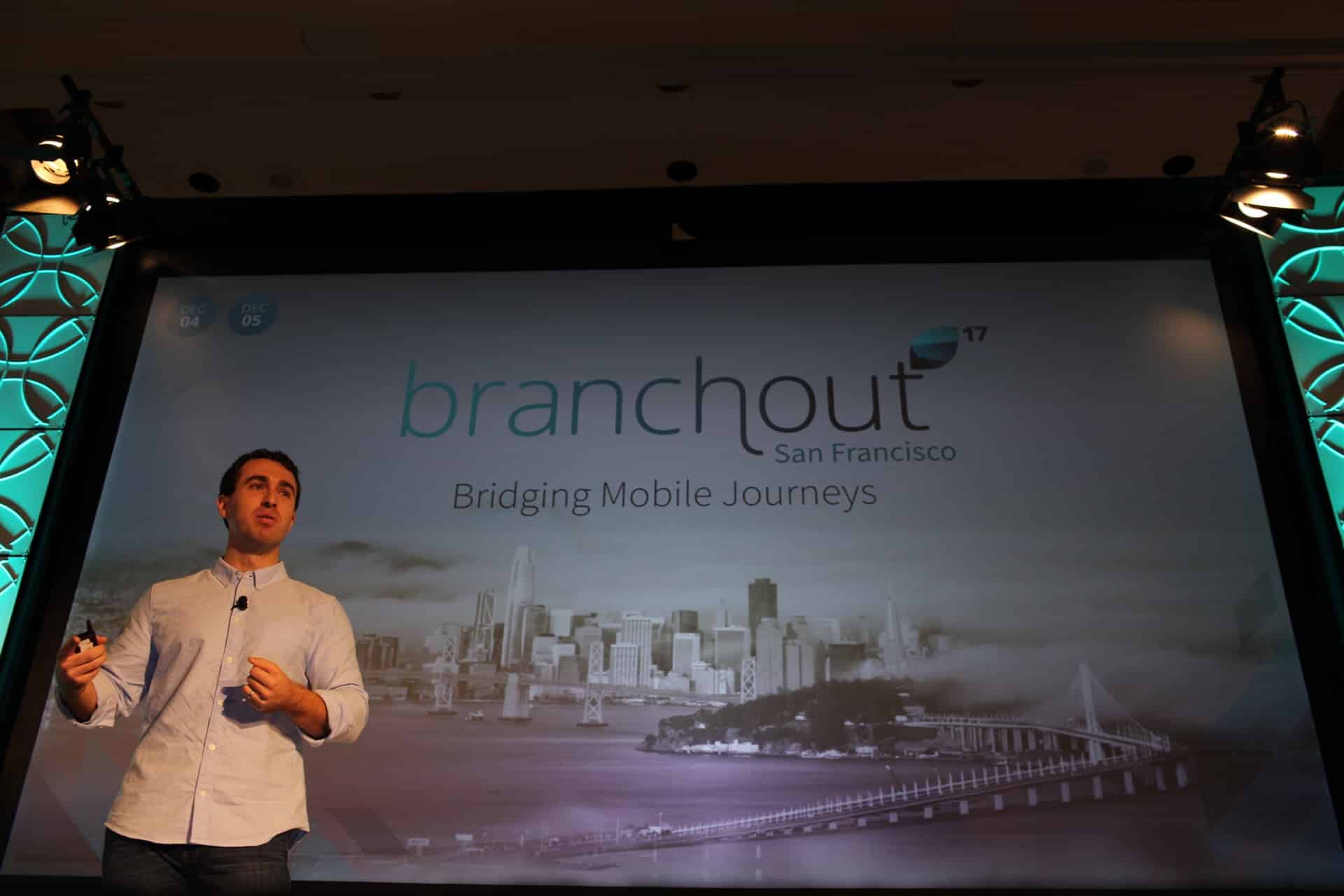 A speaker presents at the BranchOut event in San Francisco, with a backdrop showcasing the city skyline and the theme "Bridging Mobile Journeys."