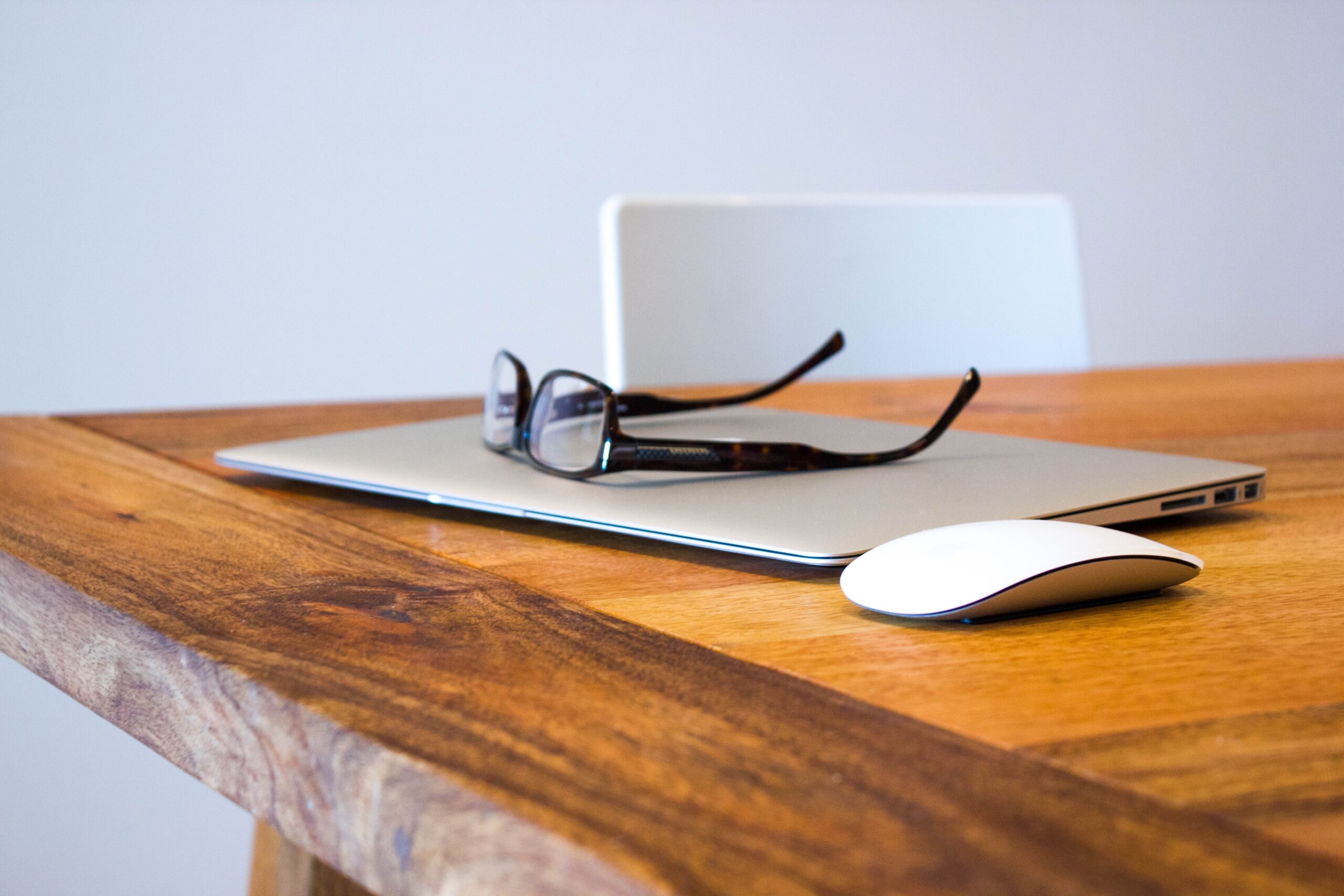 Eyeglasses on a closed laptop with a mouse on a wooden desk.