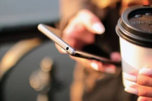 Person holding smartphone and coffee cup outdoors with sunlight.