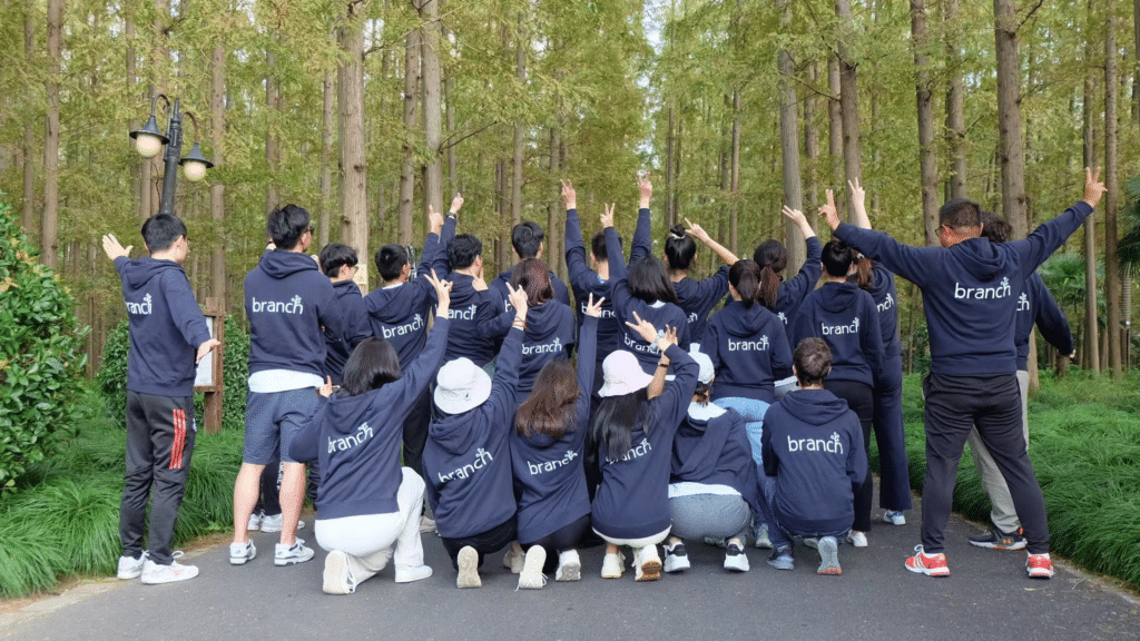 A group of Branch team members smile for a photo in a wooded area, all are wearing matching dark blue Branch sweatshirts and are facing away from the camera.