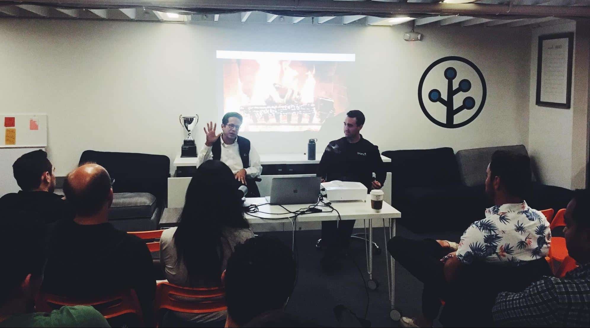 A group of attendees listens to a speaker during a presentation, with a projector displaying an image and a trophy on the table.