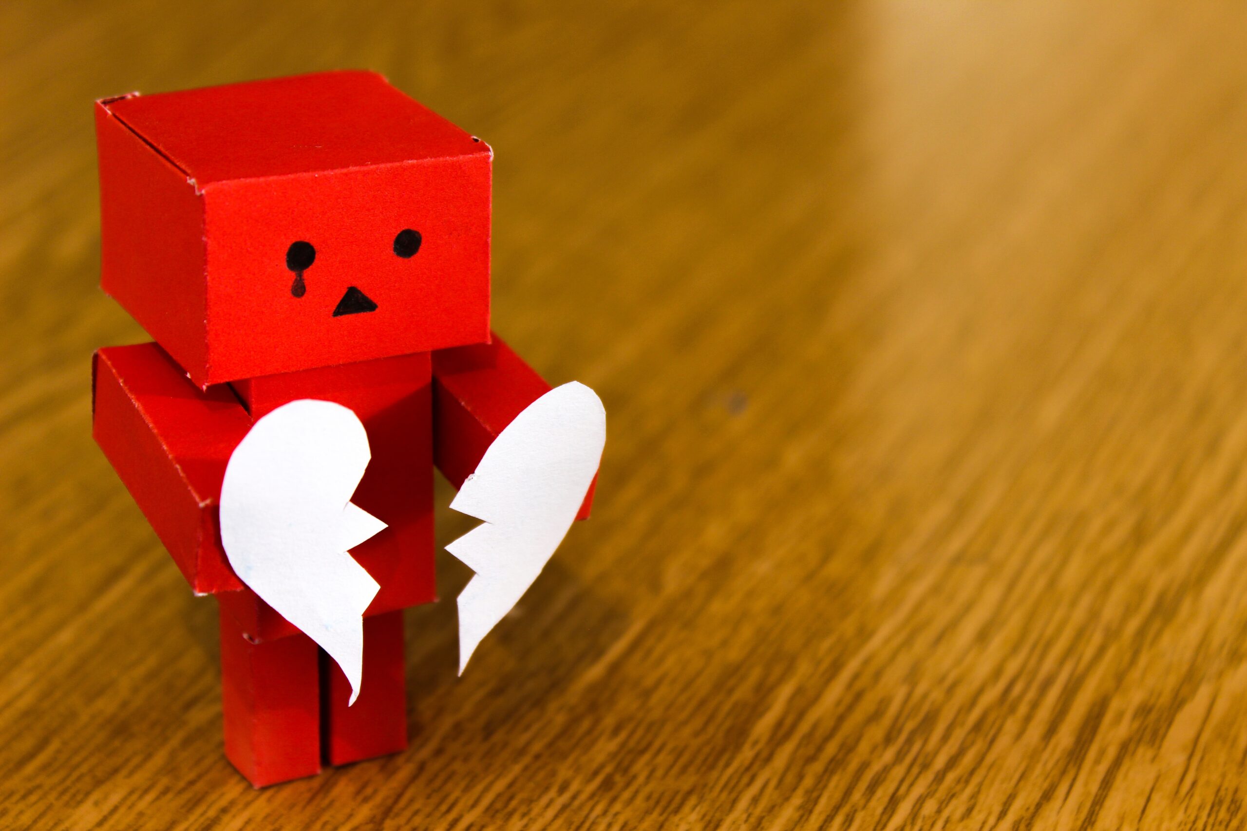 A red paper robot holding a broken white heart, expressing sadness, on a wooden surface.