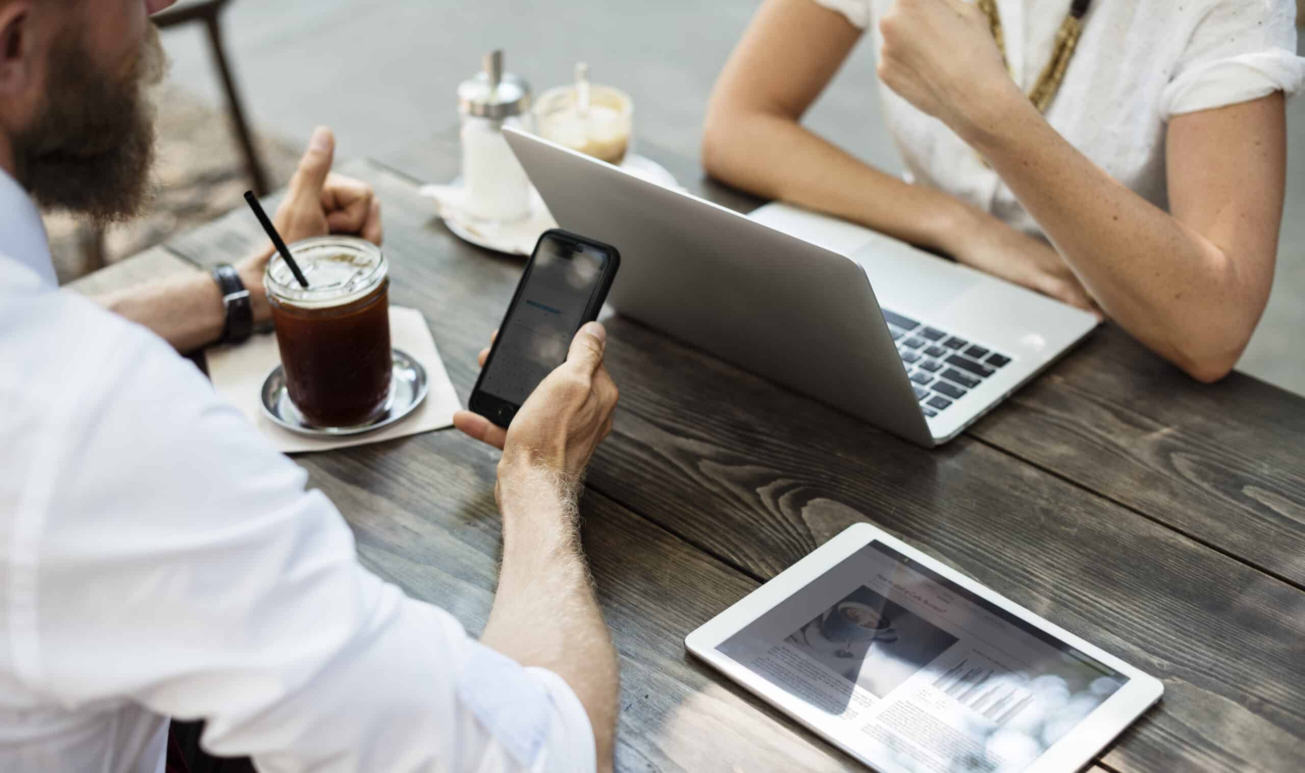 Two people at a wooden table with a laptop, mobile phone, tablet, and iced drinks.