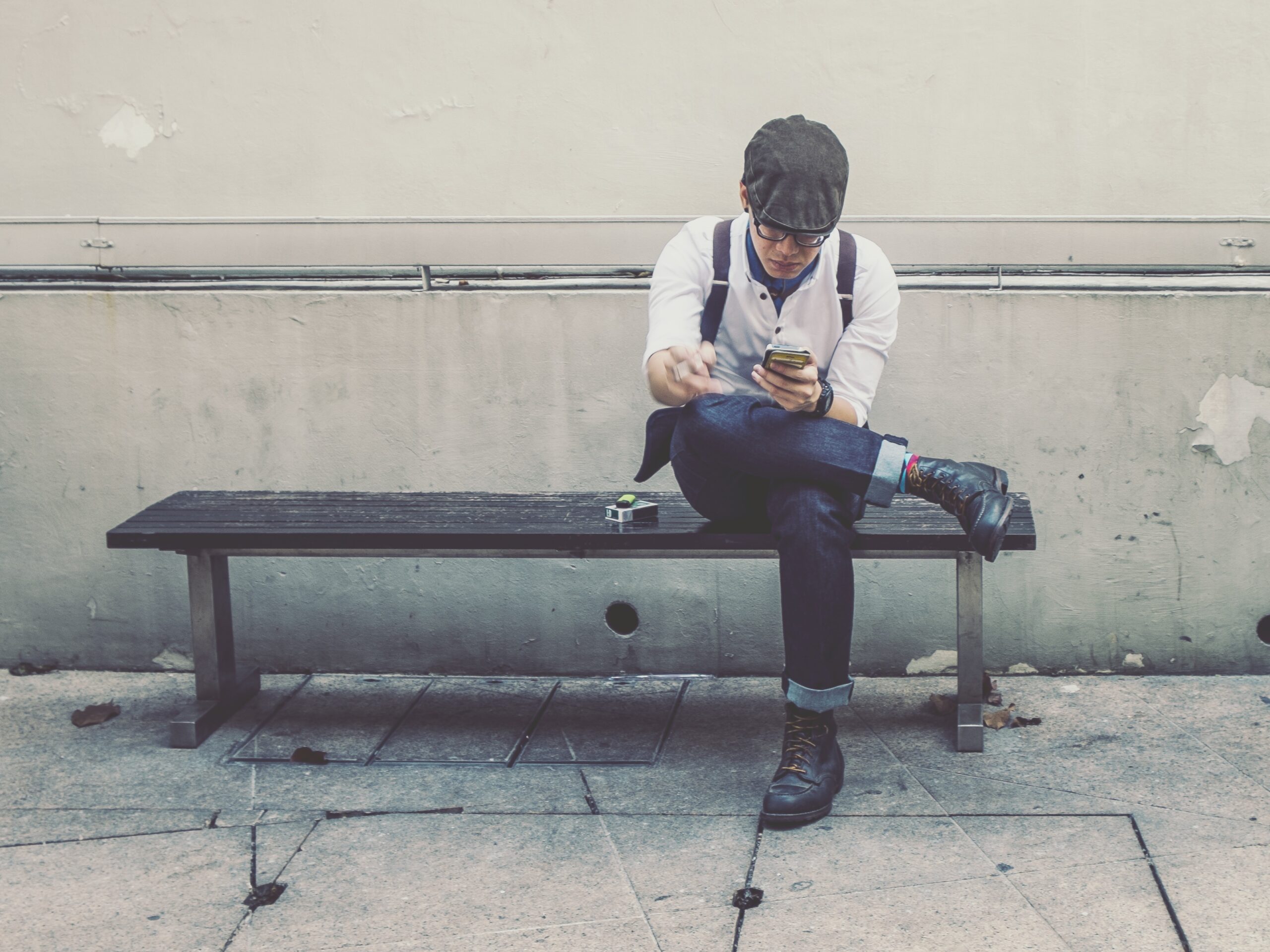 A casually dressed individual sits on a bench, focused on their smartphone with one leg crossed over the other, surrounded by urban elements.