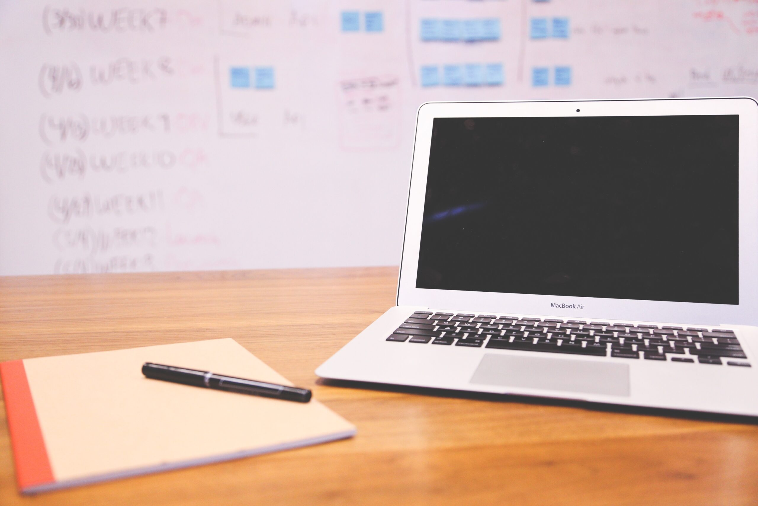 A MacBook Air on a wooden desk beside a notepad and pen, with a whiteboard covered in notes and sticky notes in the background.