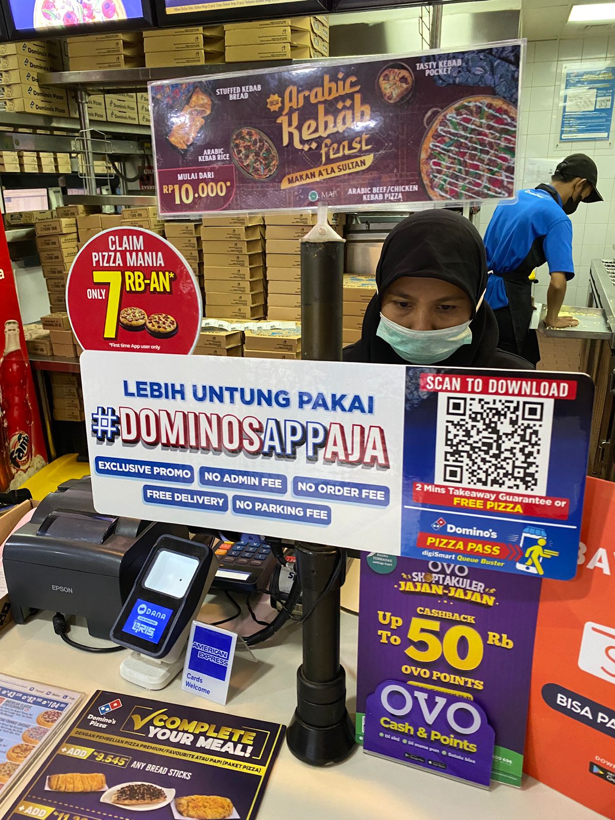 Image of an in-store cash register at Domino's showing an app promotion for the #DominosAppAja campaign. The display shows a Branch QR code that says "scan to download."