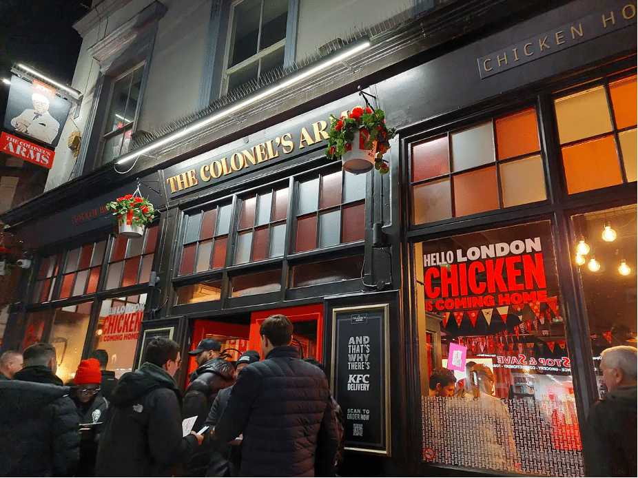 Photograph of the outside of The Colonel's Arms pub. KFC promotions are displayed in the window, including a sign that reads, "Hello London, chicken is coming home."