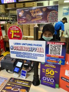 Photo of a #DominosAppAja advertisement (with QR code) in front of a register in a physical Dominos storefront.